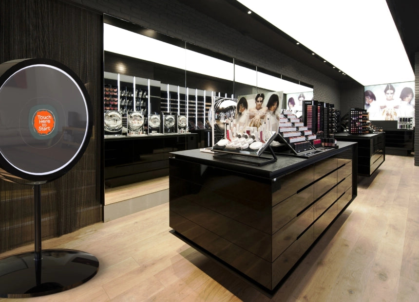Interactive photo booth with “Touch Here” display set up in modern beauty retail store featuring sleek black displays.