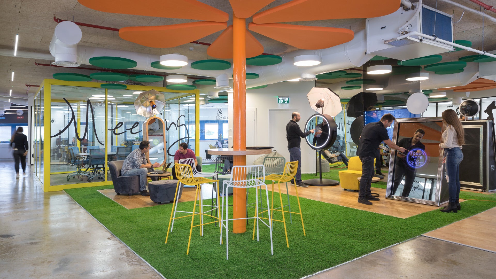 Employees at Foto Master office interacting near mirror booths and seating area, with green turf and colorful ceiling decor.