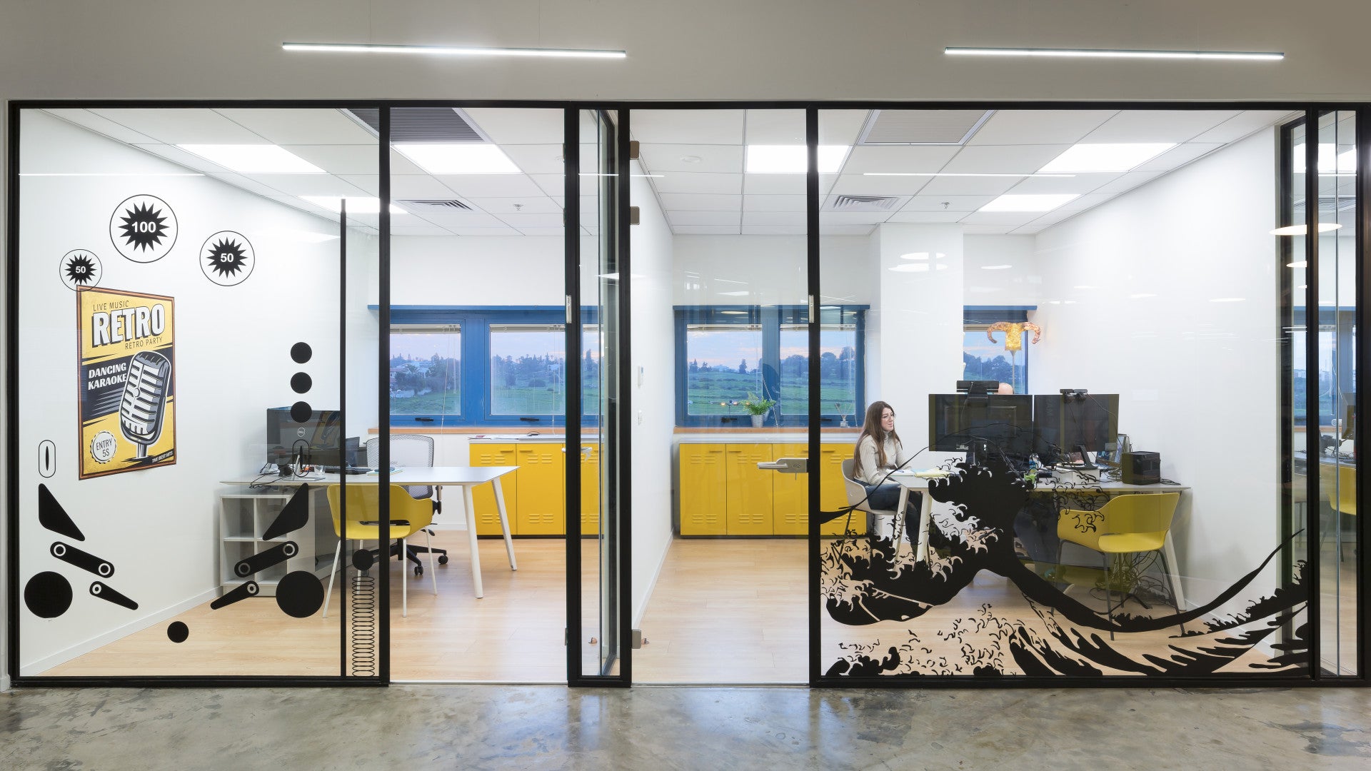 Glass-walled office at Foto Master with retro-themed wall art, yellow cabinets, and employee working at a multi-screen setup.