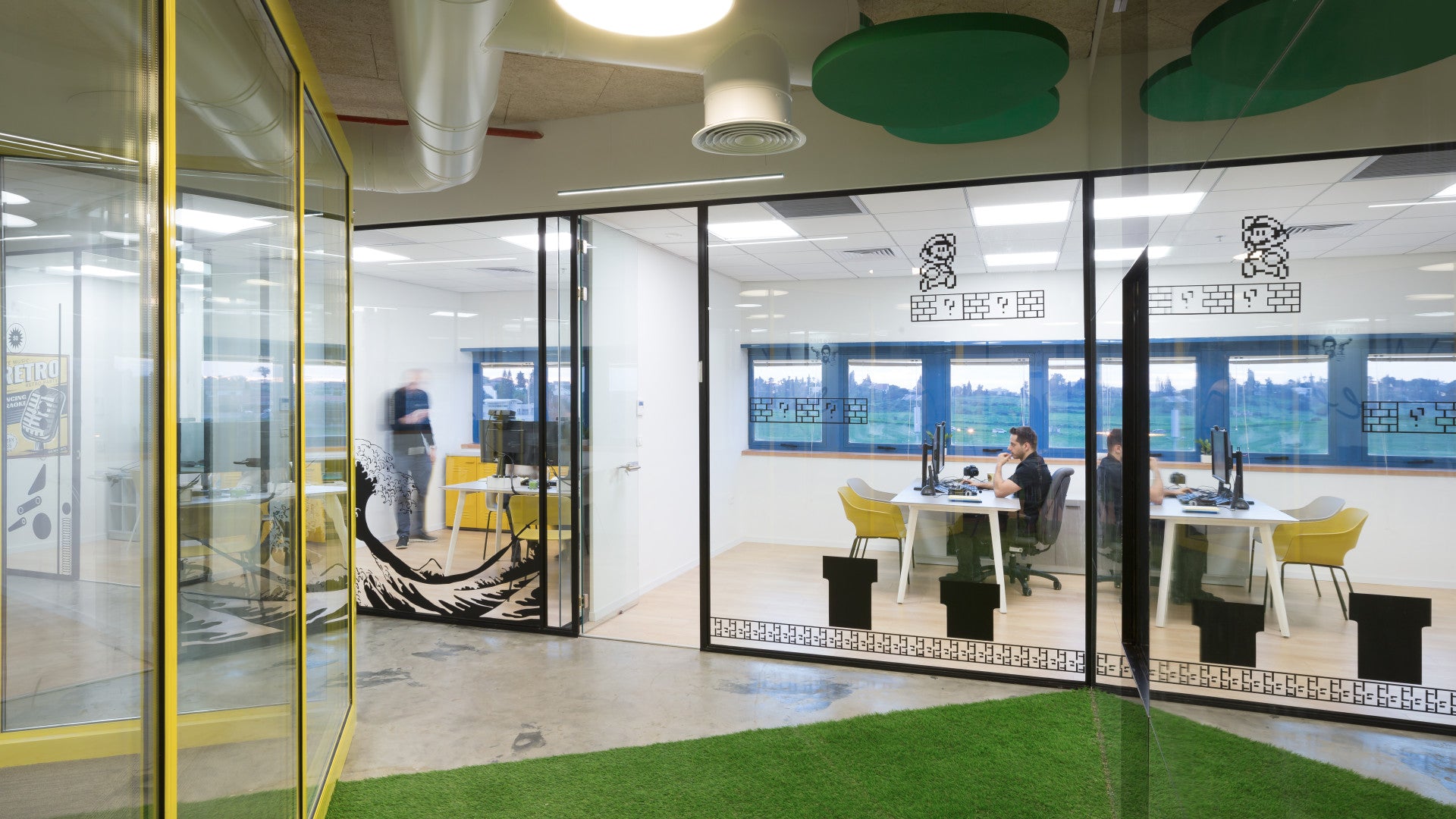 Glass-walled office at Foto Master with turf flooring, yellow chairs, gaming-themed decals, and employees at desk stations.