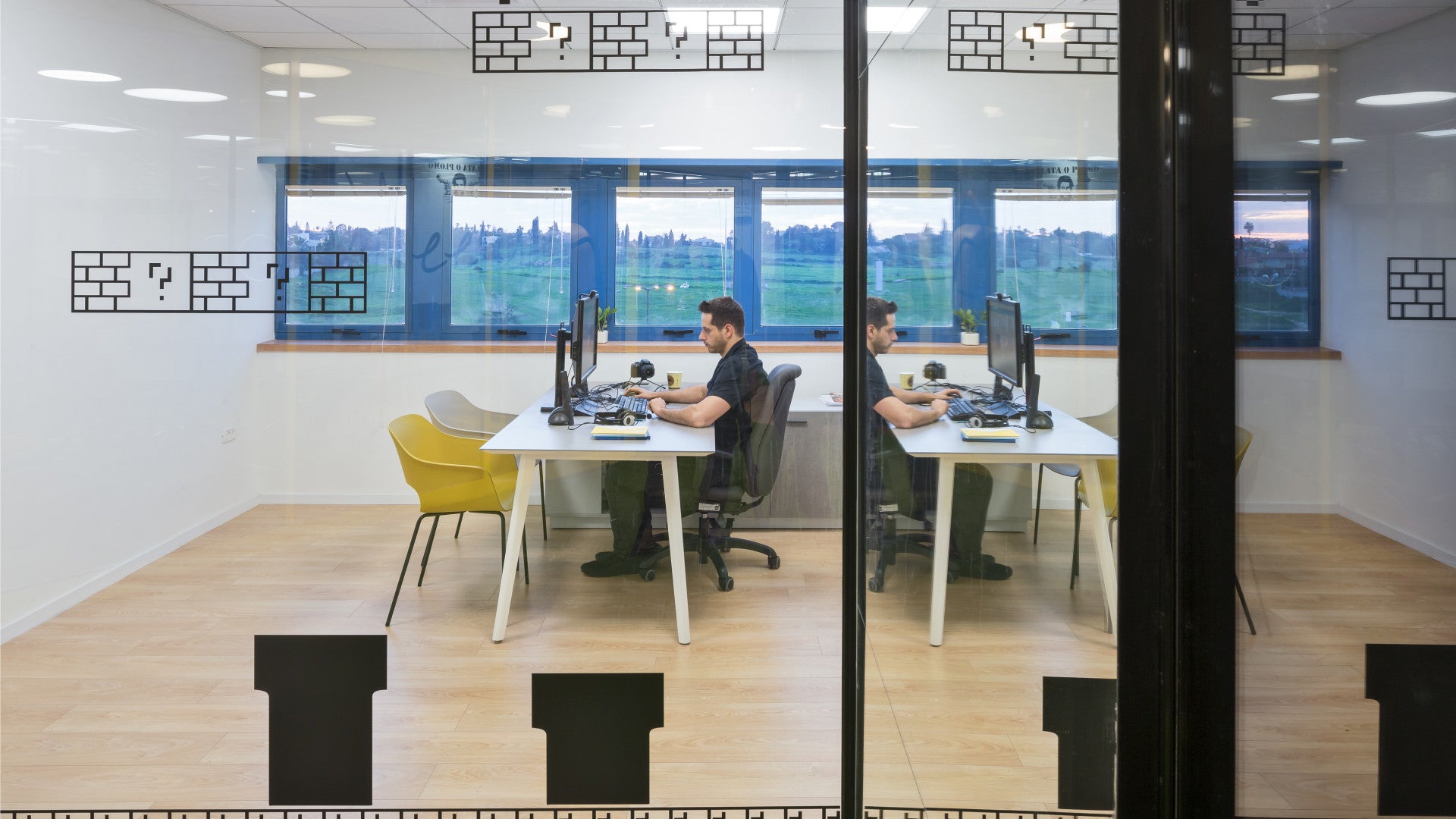 Two men working at desks with dual monitors in a glass-walled office, with large windows and wooden flooring.
