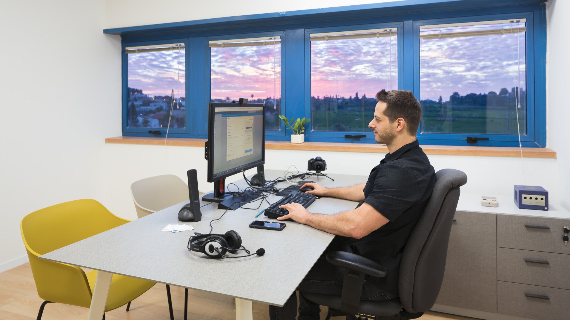 Man working at a monitor desk setup in a modern office with large windows overlooking a sunset view.